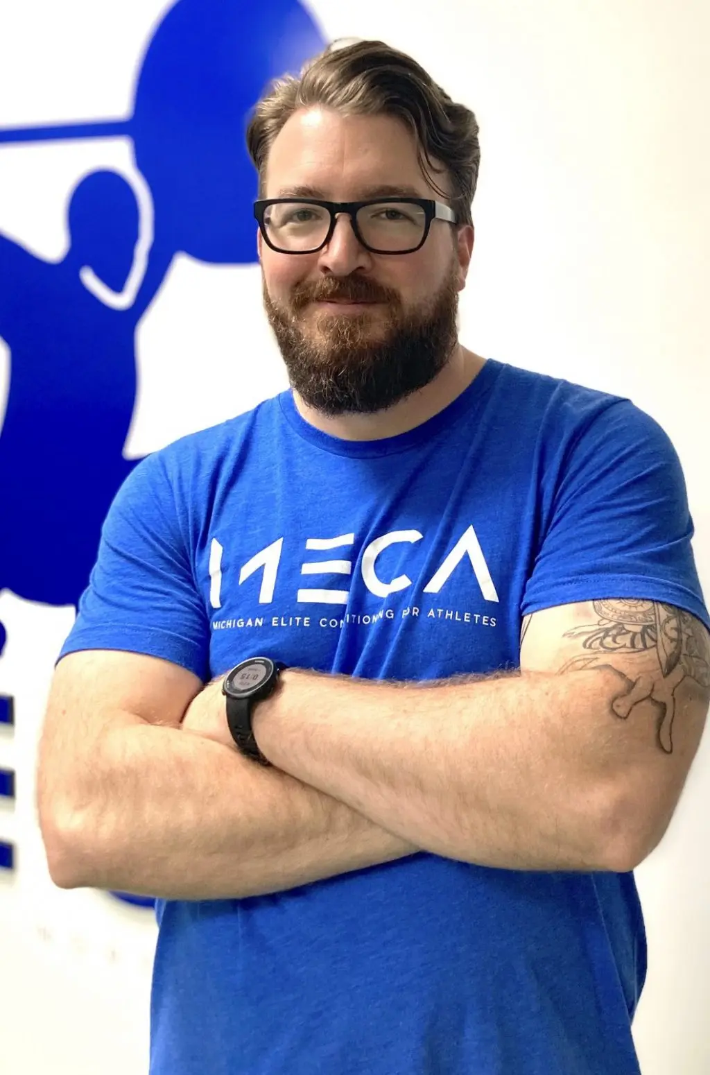 a man with a beard and glasses stands in front of a blue and white wall with a blue logo.