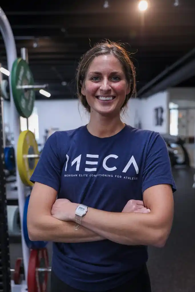 a woman in a blue shirt stands in a gym with her arms crossed.