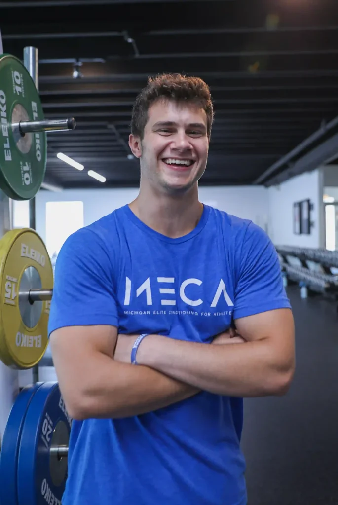 Noah Fye, an assistant strength coach at MECA Grand Rapids, poses for a headshot with gym equipment behind him.