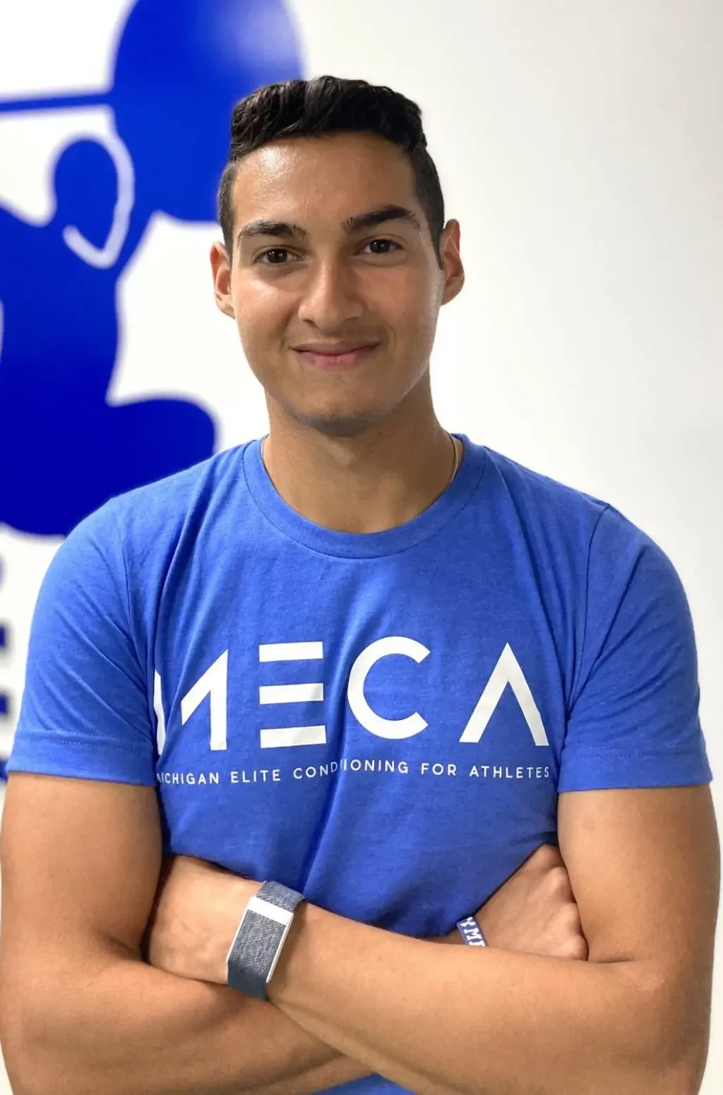 a man in a blue t - shirt stands in front of a wall with his arms crossed. His name is David and he is a assistant strength coach at MECA chicago